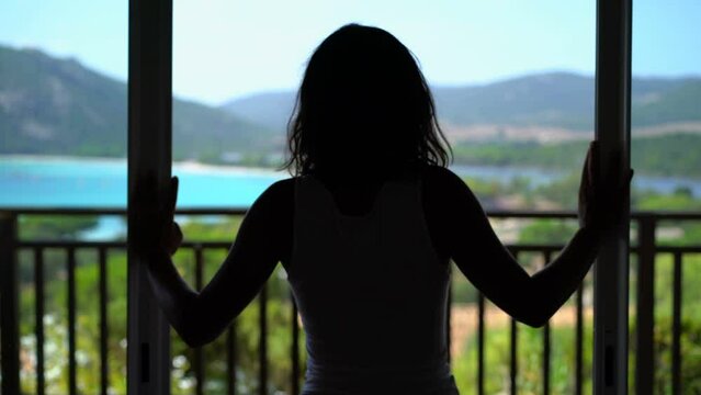 Shot of a brunette opening balcony doors and walking to a sunny terrace overlooking the sea.
