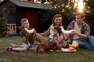 Happy young family eating melon in garden together - obrazy, fototapety, plakaty