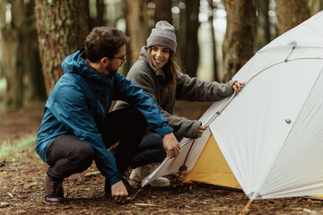 Smiling millennial caucasian guy and woman in jackets resting in cold forest, enjoy adventure, making tent - Powered by Adobe