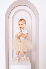 Baby girl elegant dress. A one-year-old girl in a puffy skirt and a white T-shirt poses against the backdrop of a bright room with a yellow tulip in her hand