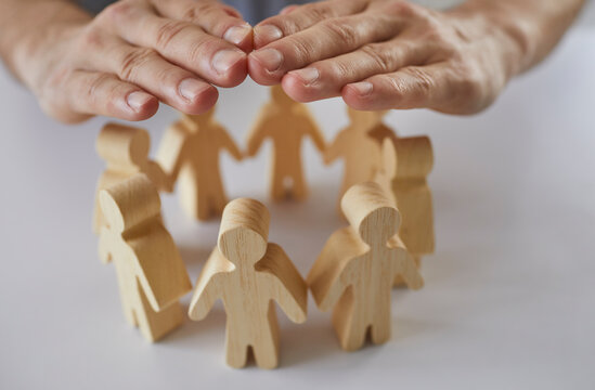Insurance agent's hands cover and protect wooden figures of people standing in circle in front of him. Close up of hands creating roof over human figurines as symbol of providing social protection.