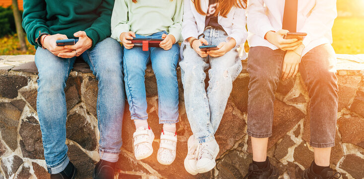 Four Kids browsing their smartphone devices while they sitting on the brick wall. Their palms with devices shot. Careless young teenhood time and a modern technology concept image.