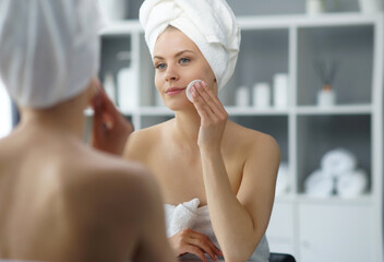 Young woman sits in the bathroom in front of the makeup mirror and does cosmetic procedures. Beautiful girl in white towel. Skin care, health, rejuvenation and spa treatment concept.