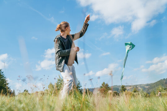 Smiling teenager boy with flying colorful kite on the high grass meadow in the mountain fields. Happy childhood moments or outdoor time spending concept image.