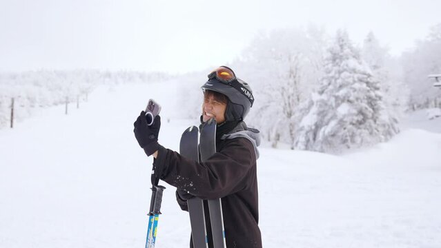 4K Young Asian man using mobile phone video call during skiing on snowy mountain at in ski resort. Handsome guy enjoy winter activity active lifestyle extreme sport training freeride ski on vacation.