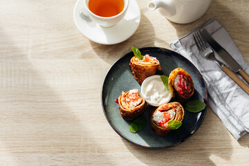 Rolls of thin pancakes with cottage cheese and sour cream, cup of tea on dark plate on wooden table. Delicious and healthy breakfast. Top view, copy space