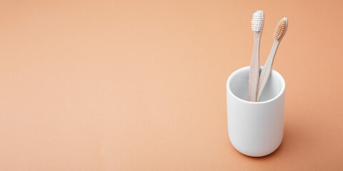 Two wooden bamboo toothbrushes in a white glass on a beige background. The concept of sustainability and the rejection of plastic. The concept of hygiene and daily routine. Copy space, web banner