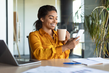 Cheerful african american female manager chatting on cellphone, sitting with cup of coffee in office interior
