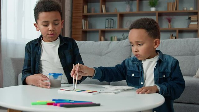 Two African American creative smart concentrate little children boys painting at home together with paints ethnic brothers kids sibling paint picture with acrylic watercolor colors art hobby education