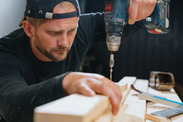 A carpenter man in dark clothes screws a self-tapping screw into a board with an electric...