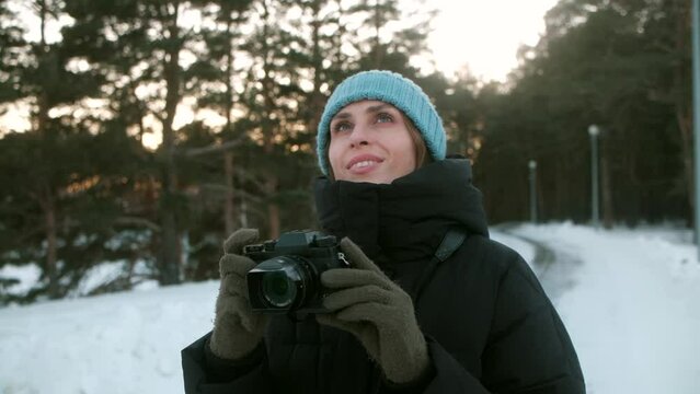 Cute young female traveler in blue hat and black jacket, take photos holding digital photo camera in hands at winter time in the park covered snow at sunset.