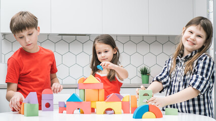 Happy school friends play toy constructor on kitchen table. Children collect colorful details of Bricks. Little kids build toy city. Home leisure, playtime. Educational interesting board game.