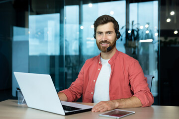 Fototapeta na wymiar Portrait of mature businessman in red shirt inside office, man with beard and video call headset smiling and looking at camera, online customer support worker with laptop.