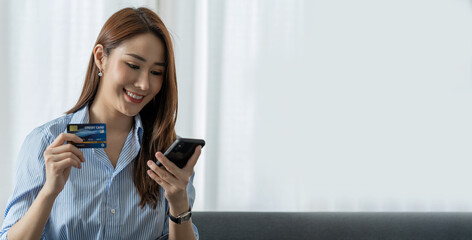 Cute and bright Asian businesswoman shopping online on mobile comfortably using a credit card to purchase goods for the convenience of spending at home on weekends.