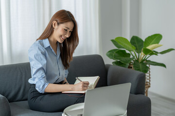 Confident Asian businesswoman sitting and taking notes in financial book Income tax with laptop computer or tablet in a happy office.