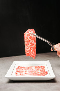 Premium Rare Slices sirloin Wagyu A5 beef with high-marbling texture on a ceramic plate, pick up by bbq tongs with hand. Served for Yakiniku, Sukiyaki and Shabu. Image with copy space.