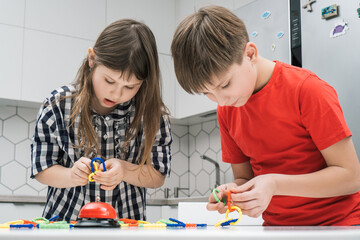 Cute school kids play toy constructor standing above kitchen table and looking down. Boy and girl collect plastic colored details of blocks. Home leisure, playtime. Entertaining interesting board game.