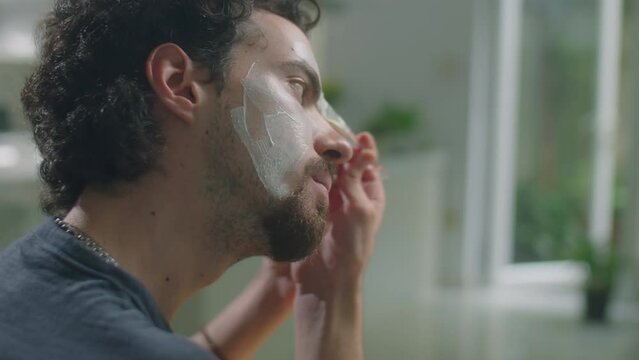 Rack focus shot of male beauty influencer applying clay mask to face with cosmetic brush in front of circle lamp and smartphone while filming vlog at home