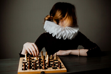 A beautiful woman in a black dress with a white jabot is sitting at a table at the chessboard with...