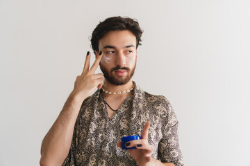 Young latin gay queer man applying a facial cream to take care of his skin on a white background.