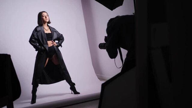 sexy young fashionable woman model in a black swimsuit and leather boots and coat is photographed in the studio with a photographer on a white background. backstage