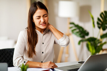 Young woman is sitting at a desk in the office and has neck pain while working with a laptop - 571584312