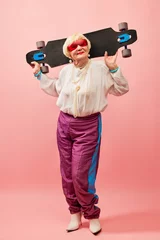 Afwasbaar fotobehang Energetic lady. Beautiful old woman, grandmother in stylish clothes posing with skateboard over pink studio background. Concept of age, fashion, lifestyle, emotions, facial expression © master1305