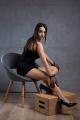 Classic dark studio portrait of a young brunette woman in black clothes who is sitting on a chair against a concrete wall.