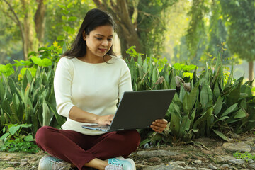 Happy Indian girl working on a Laptop in a lush green park in day time in summer. Busy with work he...