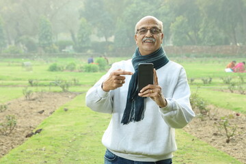portrait of happy Indian old man in green park showing mobile phone doing thumbs up.