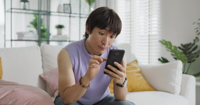 Adult people asia single LGBT gay man sitting at home sofa looking at dating game tinder app on mobile phone. Relax smile choose lover enjoy playing right swipe to find love couple match flirt chat.