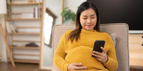 Happy Asian pregnant woman using her phone while relaxing on sofa in her living room