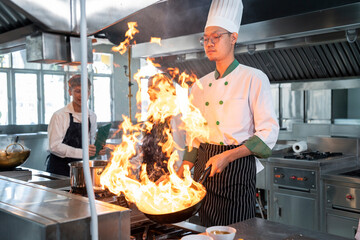 Asian male chef stir-frying vegetables over a blazing fire for Chinese stir-fry fresh vegetables in a restaurant