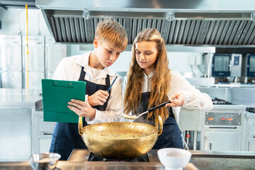 Teenager male female students learn to cook in class in the kitchen Teenager male holding clipboard taking recipe notes Cooking female teenage student practicing making desserts on the kitchen stove