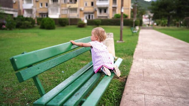 Little girl climbs onto a bench and stands on her feet, holding on to the back