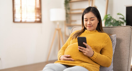 Fototapeta na wymiar Happy Asian pregnant woman using her phone while relaxing on sofa in her living room