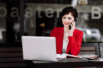 Smiling businesswoman talking on the smartphone, working on a laptop and doing notes at cafe on terrace in urban city. Office worker communication with clients remote. Coffee break, business concept