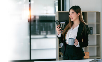 Smiling businesswoman in suit using mobile phone standing in open office.