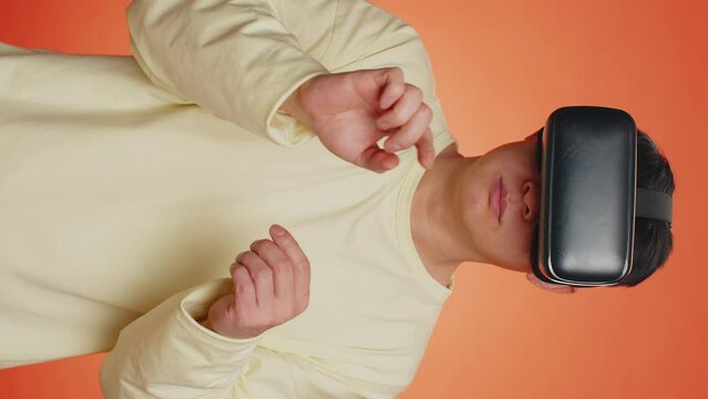 Asian man using headset helmet to play simulation game app online. Watching virtual reality 3D 360 video content. Guy in VR goggles isolated on orange background. Futuristic technology. Vertical shot