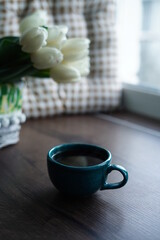 A close-up of a turquoise cup of black coffee against a basket of white tulips. A beautiful blue cup of hot coffee sits on a wooden table near the window