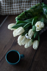 A top view of a bouquet of white tulips and a turquoise cup of black coffee. A beautiful blue cup with hot coffee and a bouquet of white tulips stands on a wooden table near the window