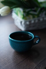A close-up of a turquoise cup of black coffee against a basket of white tulips. A beautiful blue cup of hot coffee sits on a wooden table near the window