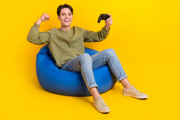 Full body portrait of delighted cheerful man sit comfort bag raise fist isolated on yellow color background