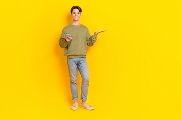 Fototapeta na wymiar Full body photo of positive young man hold smart phone arm palm demonstrate empty space isolated on yellow color background