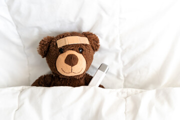 Ein kleiner brauner Teddybär mit einem Fieberthermometer und einem Heftpflaster in einem Bett.