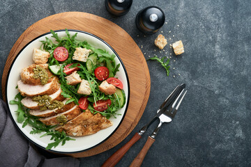 Fresh salad with grilled chicken fillet, lettuce, arugula, spinach, tomatoes, cucumber and pesto sauce, sesame seeds with olive oil in white bowl on black slate background. Healthy lunch menu Top view