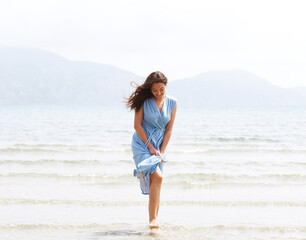 Fototapeta na wymiar Young brunette woman in a blue dress walking barefoot on a beach and dangles his feet in the water.