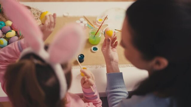 Top view of woman and her little daughter in rabbit ears decorating Easter eggs