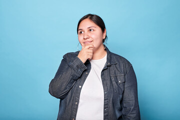 Young curvy latina woman looks empty space, hand on chin, wearing denim shirt and hoop earrings and...