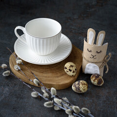 Festive Easter spring composition with white ceramic tea pair and quail eggs on wooden podium with willow branches and handmade paper rabbit on dark background